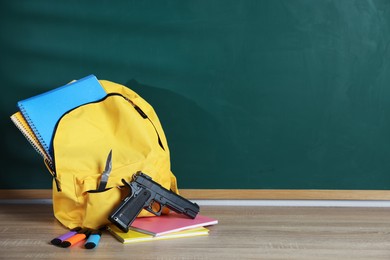 Photo of Backpack with knife, gun and stationery on wooden table near blackboard, space for text