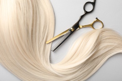 Photo of Blonde hair lock and scissors on light grey background, top view