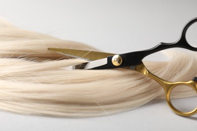 Photo of Cutting blonde hair lock with scissors on light grey background, closeup