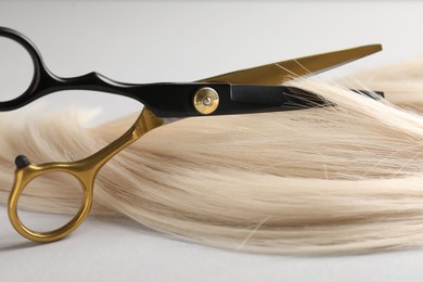 Photo of Cutting blonde hair lock with scissors on light grey background, closeup