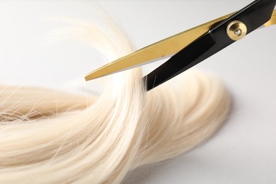 Photo of Cutting blonde hair lock with scissors on light grey background, closeup
