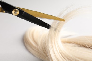 Photo of Cutting blonde hair lock with scissors on light grey background, closeup