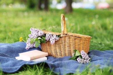 Photo of Stacked books, blanket and wicker basket with lilac flowers on green grass outdoors, closeup