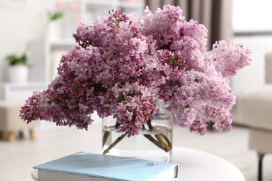 Photo of Beautiful lilac flowers in vase and book on table indoors, closeup