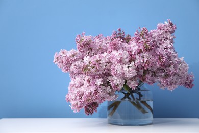 Photo of Beautiful lilac flowers in glass vase on white table near light blue wall, space for text