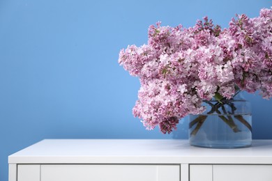 Photo of Beautiful lilac flowers in glass vase on cabinet near light blue wall, space for text