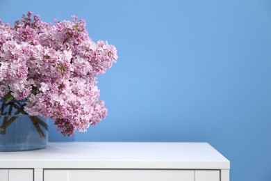 Photo of Beautiful lilac flowers in glass vase on cabinet near light blue wall, space for text