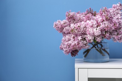 Photo of Beautiful lilac flowers in glass vase on cabinet near light blue wall, space for text