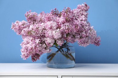 Photo of Beautiful lilac flowers in glass vase on cabinet near light blue wall