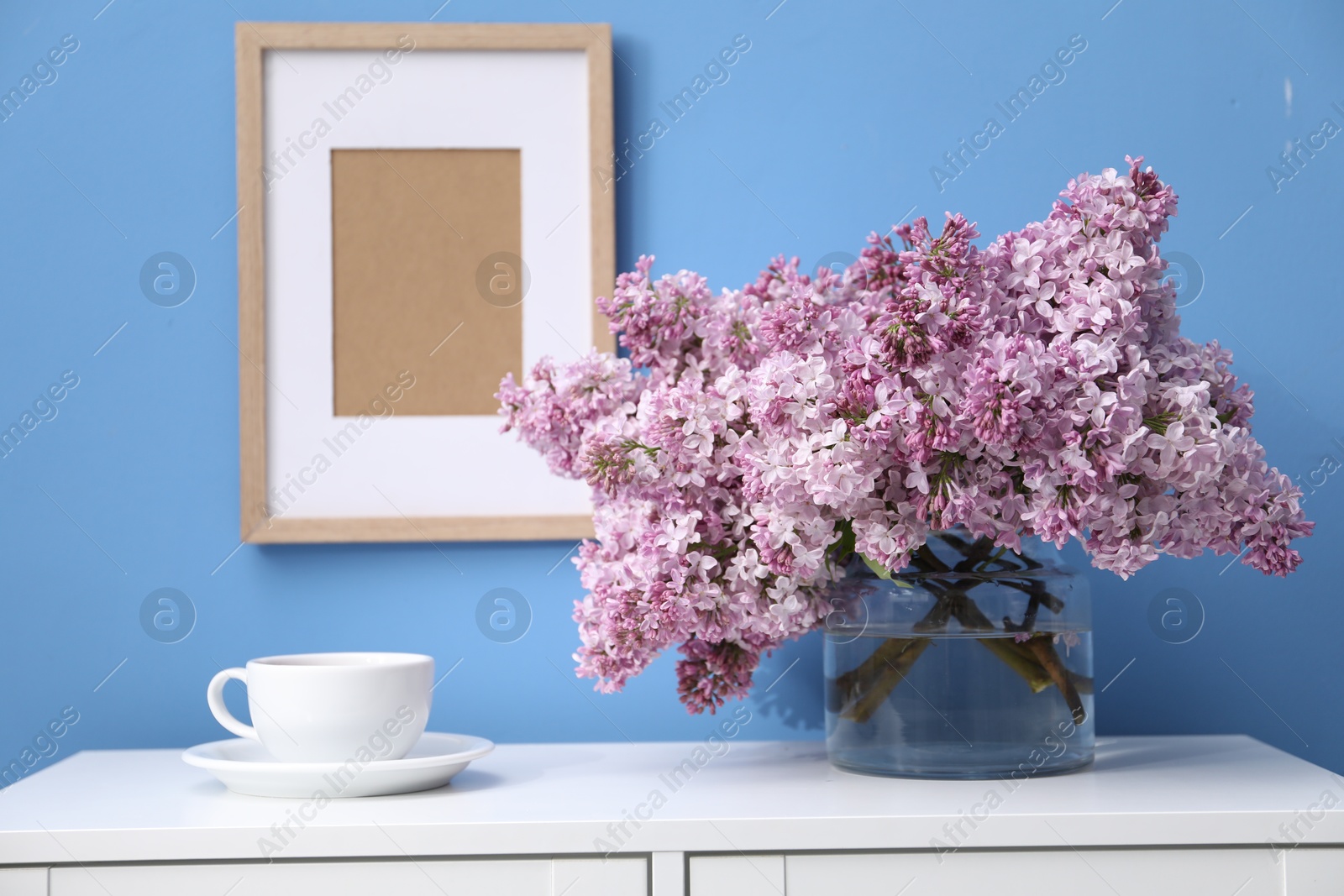 Beautiful lilac flowers in glass vase and cup on cabinet near light blue wall Photo of Beautiful lilac flowers in glass vase and cup on cabinet near light blue wall