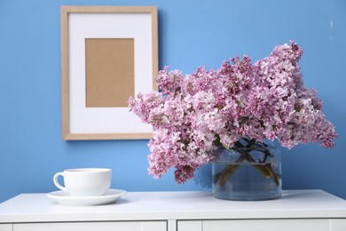 Photo of Beautiful lilac flowers in glass vase and cup on cabinet near light blue wall