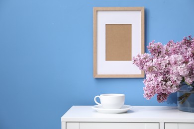 Photo of Beautiful lilac flowers in glass vase and cup on cabinet near light blue wall, space for text