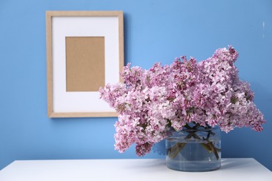 Photo of Beautiful lilac flowers in glass vase on cabinet near light blue wall and picture frame indoors