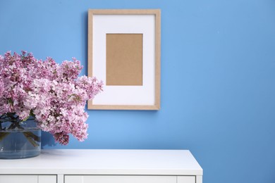 Photo of Beautiful lilac flowers in glass vase on cabinet near light blue wall and picture frame indoors, space for text
