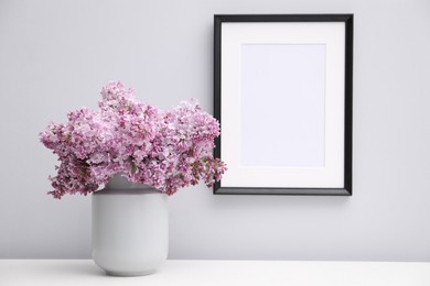 Photo of Beautiful lilac flowers in glass vase on table near white wall and picture frame indoors