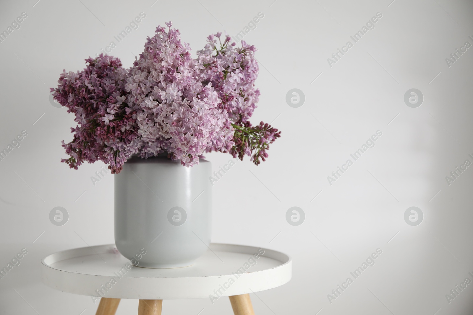 Beautiful lilac flowers in vase on table near white wall indoors, space for text Photo of Beautiful lilac flowers in vase on table near white wall indoors, space for text