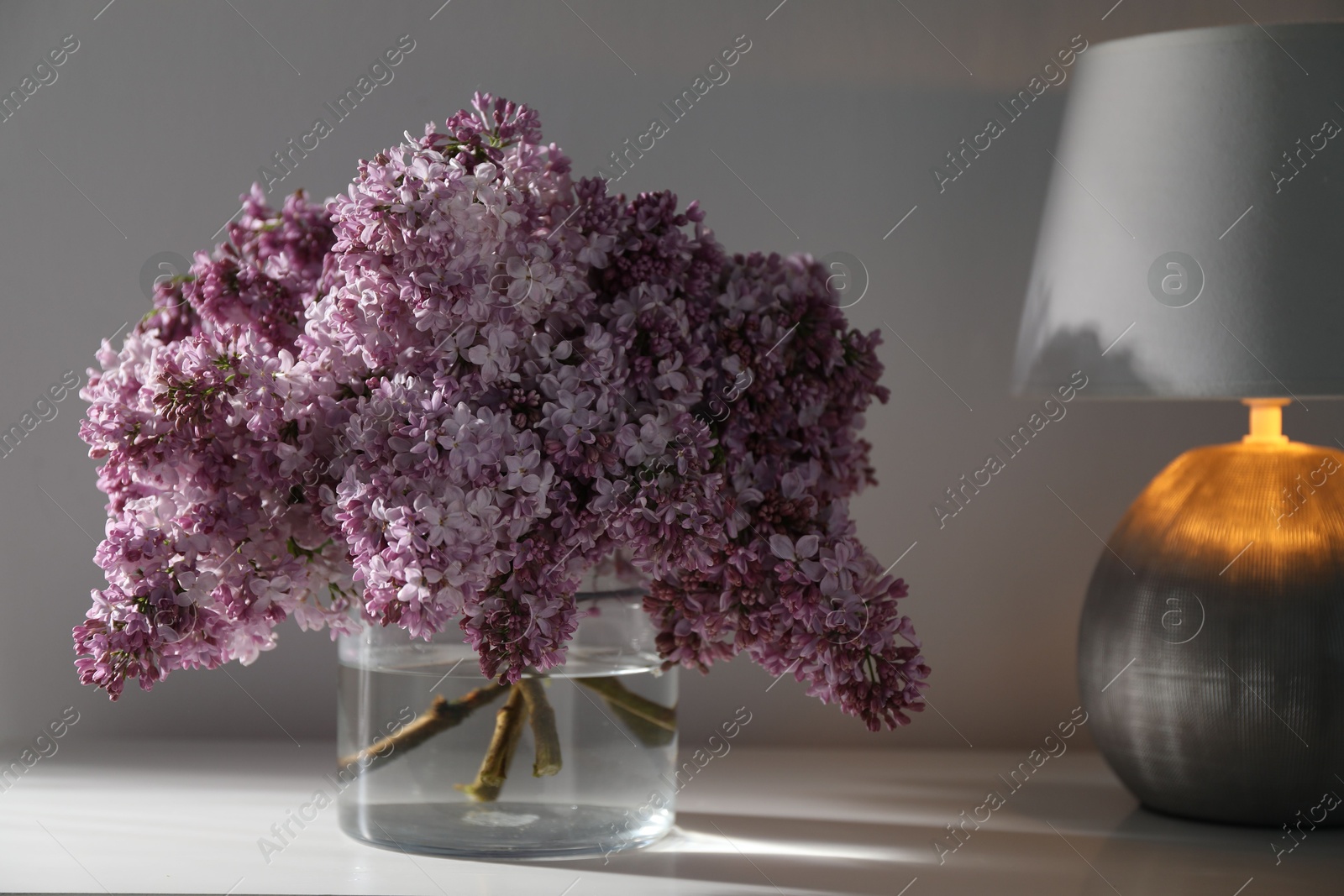 Beautiful lilac flowers in glass vase and lamp on cabinet near white wall indoors Photo of Beautiful lilac flowers in glass vase and lamp on cabinet near white wall indoors