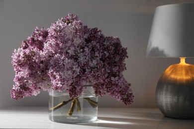 Photo of Beautiful lilac flowers in glass vase and lamp on cabinet near white wall indoors