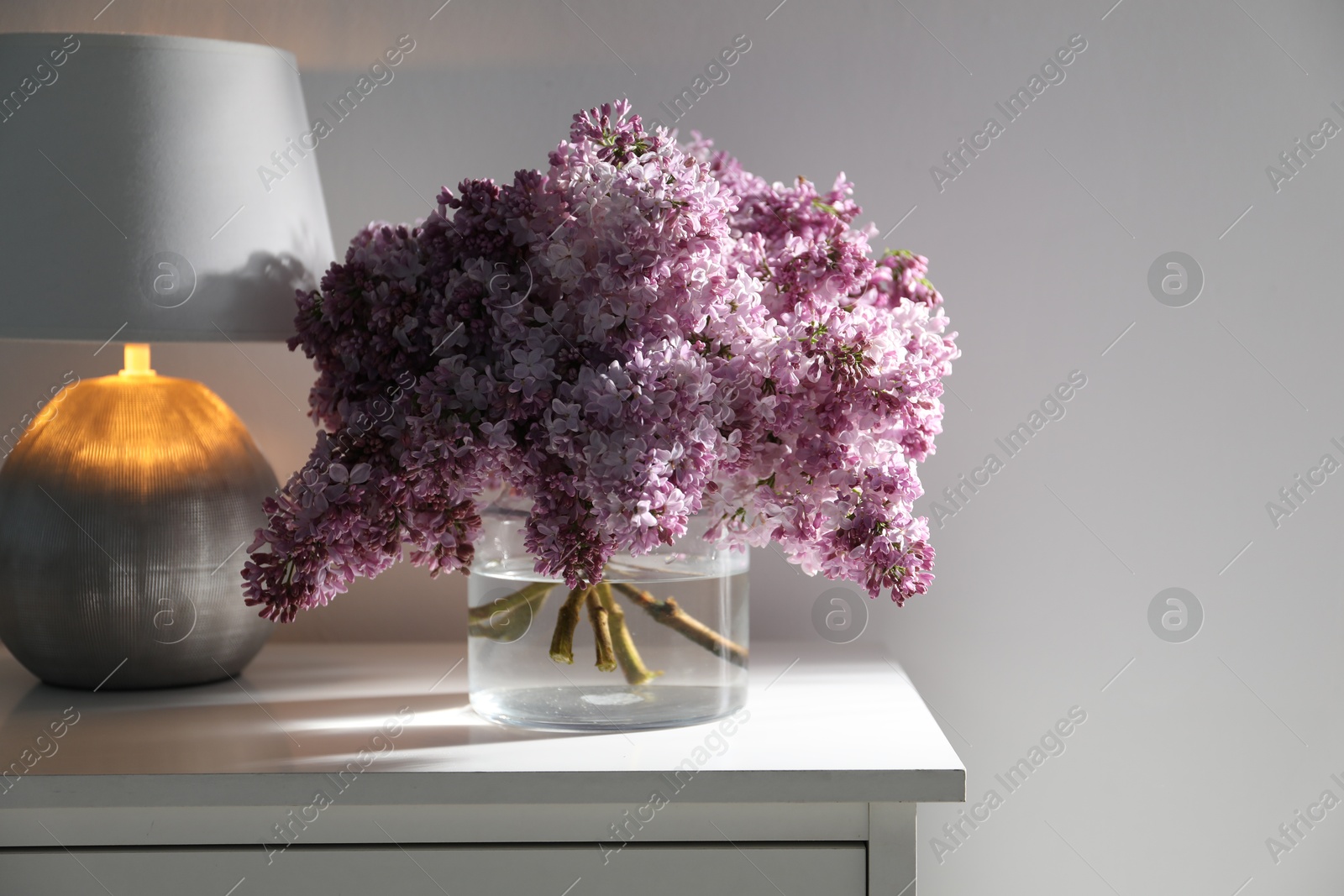 Beautiful lilac flowers in glass vase and lamp on cabinet near white wall indoors, space for text Photo of Beautiful lilac flowers in glass vase and lamp on cabinet near white wall indoors, space for text