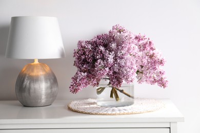 Photo of Beautiful lilac flowers in glass vase and lamp on cabinet near white wall indoors