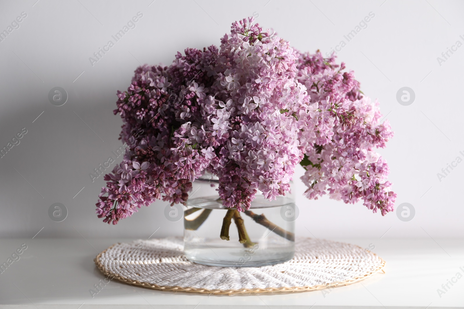 Beautiful lilac flowers in glass vase on table near white wall indoors Photo of Beautiful lilac flowers in glass vase on table near white wall indoors