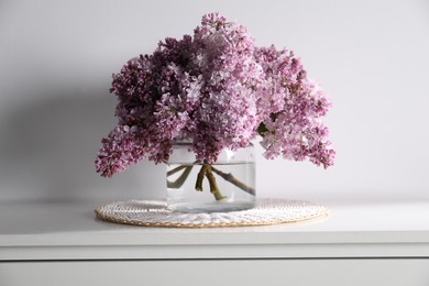 Photo of Beautiful lilac flowers in glass vase on cabinet near white wall indoors