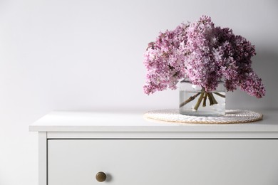 Photo of Beautiful lilac flowers in glass vase on cabinet near white wall indoors. Space for text