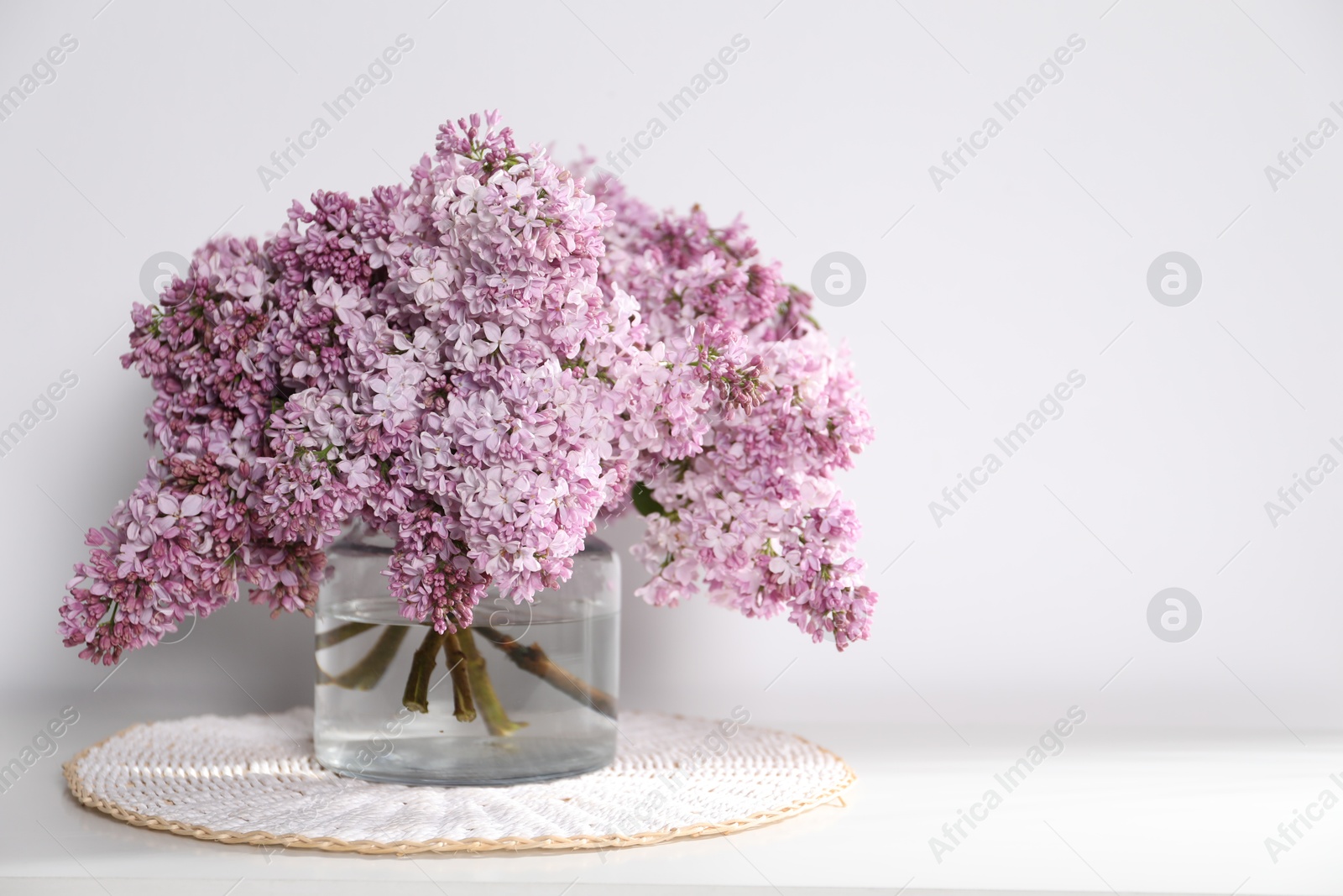 Beautiful lilac flowers in glass vase on table near white wall, space for text Photo of Beautiful lilac flowers in glass vase on table near white wall, space for text