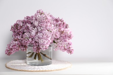 Photo of Beautiful lilac flowers in glass vase on table near white wall, space for text