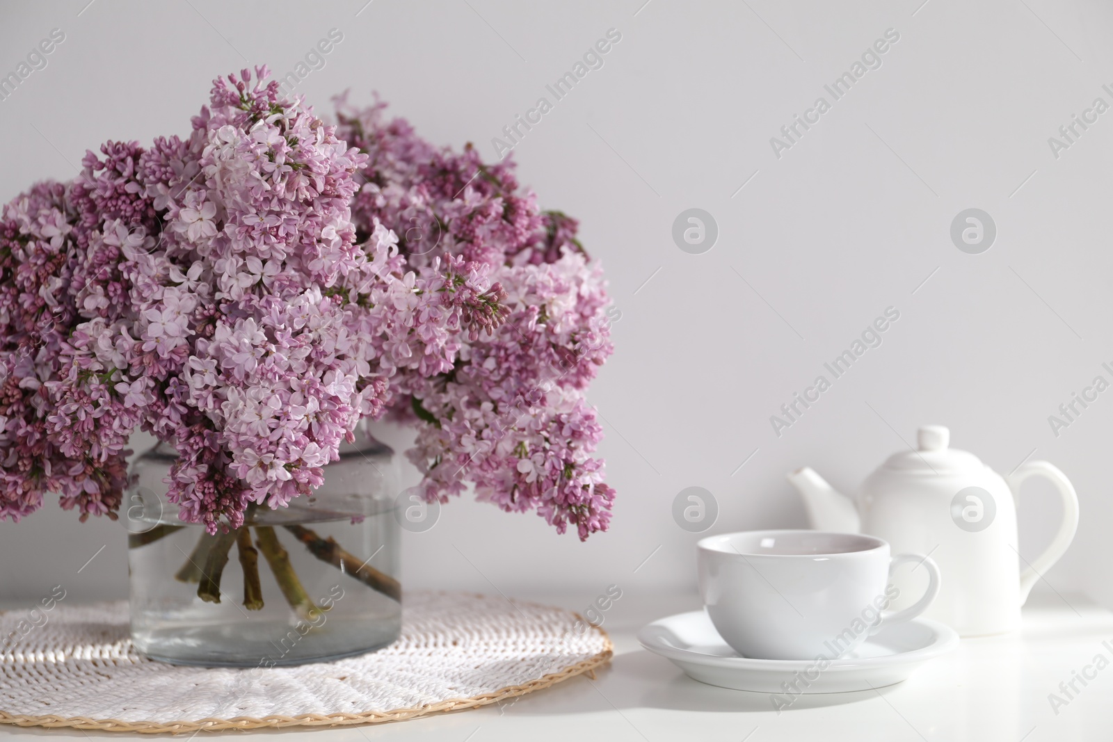Beautiful lilac flowers in glass vase, cup of drink and teapot on table near white wall indoors Photo of Beautiful lilac flowers in glass vase, cup of drink and teapot on table near white wall indoors