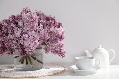 Photo of Beautiful lilac flowers in glass vase, cup of drink and teapot on table near white wall indoors