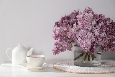 Photo of Beautiful lilac flowers in glass vase, cup of drink and teapot on table near white wall indoors