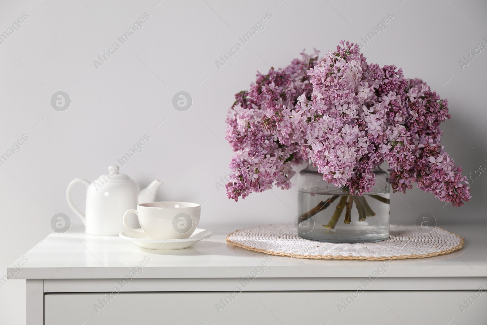 Beautiful lilac flowers in glass vase, cup of drink and teapot on cabinet near white wall indoors Photo of Beautiful lilac flowers in glass vase, cup of drink and teapot on cabinet near white wall indoors