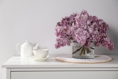 Photo of Beautiful lilac flowers in glass vase, cup of drink and teapot on cabinet near white wall indoors