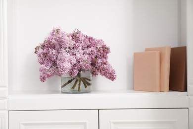 Photo of Beautiful lilac flowers in glass vase and books on shelf indoors