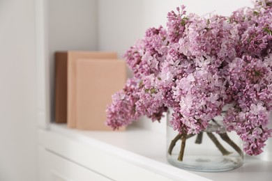 Photo of Beautiful lilac flowers in glass vase and books on shelf indoors, closeup. Space for text