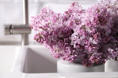 Photo of Beautiful lilac flowers in kitchen sink indoors, closeup