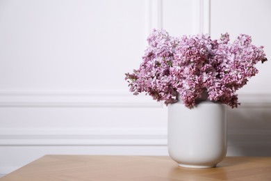 Photo of Beautiful lilac flowers in vase on wooden table near white wall indoors, space for text