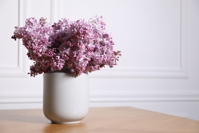 Photo of Beautiful lilac flowers in vase on wooden table near white wall indoors, space for text
