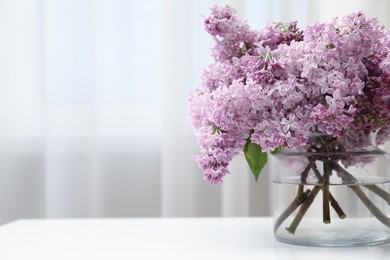 Photo of Beautiful lilac flowers in glass vase on white table indoors, closeup. Space for text