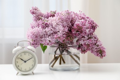 Photo of Beautiful lilac flowers in glass vase and alarm clock on table indoors