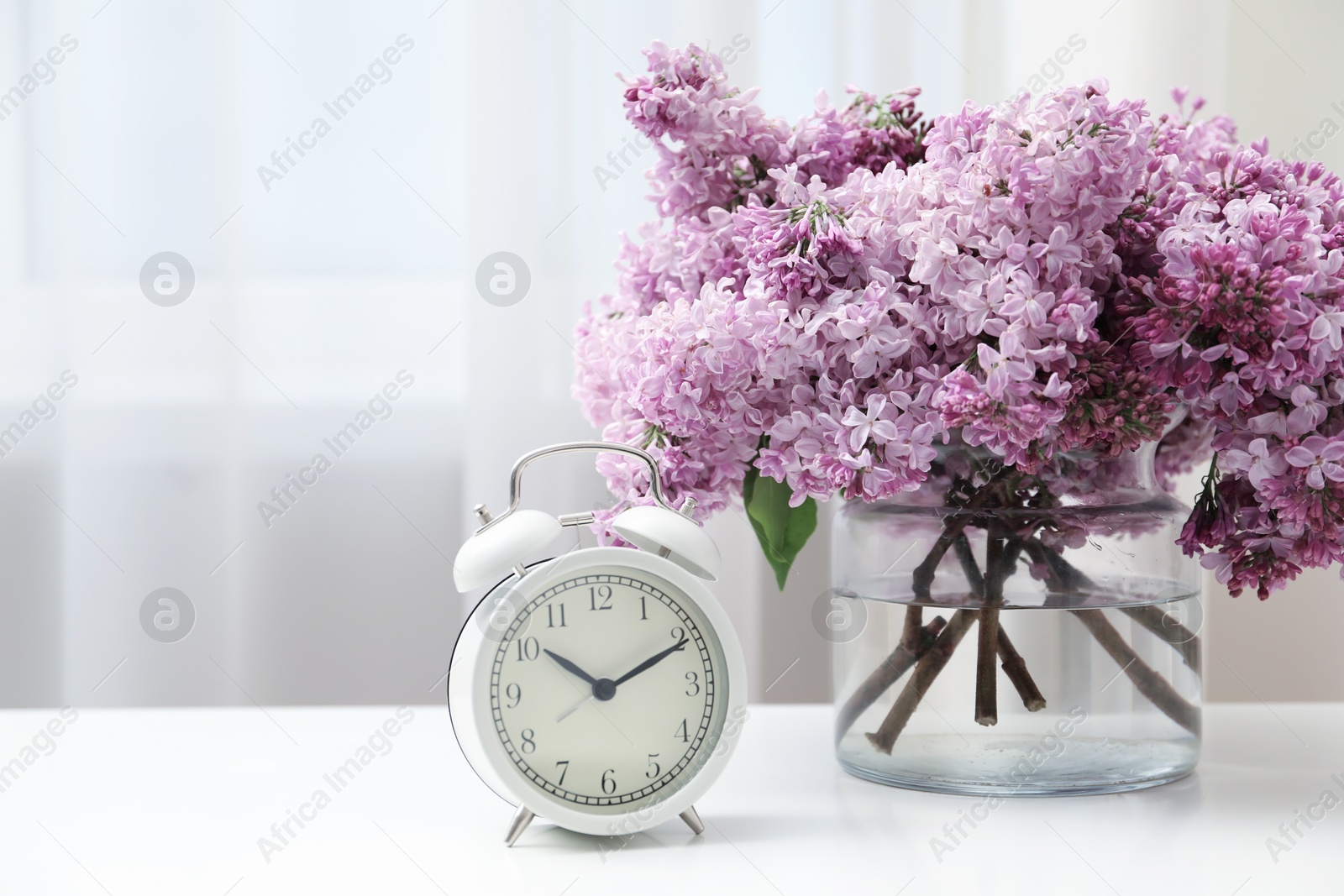 Beautiful lilac flowers in glass vase and alarm clock on table indoors Photo of Beautiful lilac flowers in glass vase and alarm clock on table indoors