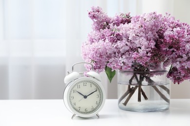 Photo of Beautiful lilac flowers in glass vase and alarm clock on table indoors