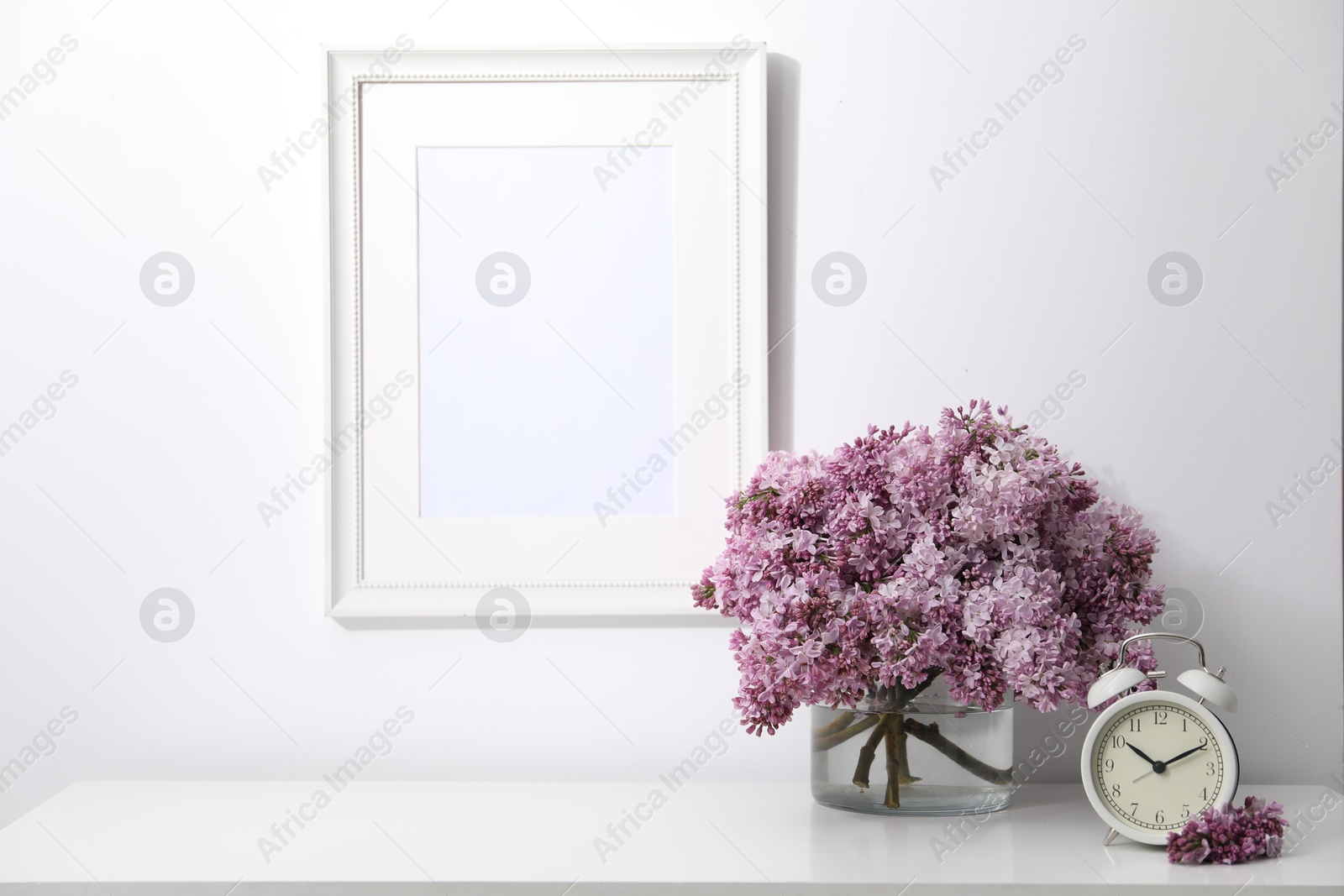 Beautiful lilac flowers and alarm clock on table near white wall indoors Photo of Beautiful lilac flowers and alarm clock on table near white wall indoors