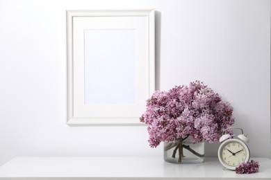 Photo of Beautiful lilac flowers and alarm clock on table near white wall indoors