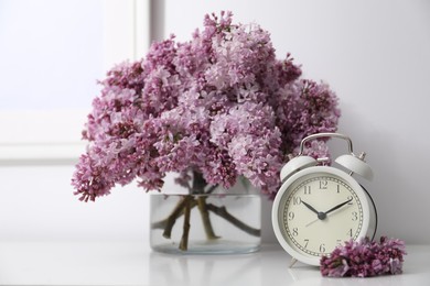 Photo of Beautiful lilac flowers and alarm clock on table near white wall indoors