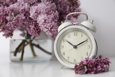 Photo of Beautiful lilac flowers and alarm clock on table near white wall indoors, closeup