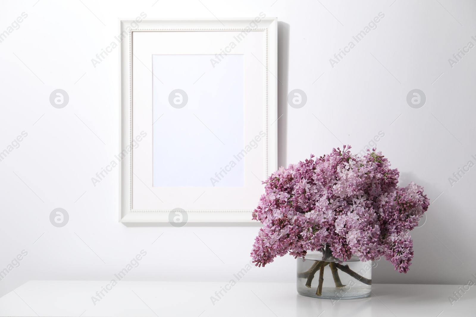 Beautiful lilac flowers in glass vase on table and picture frame indoors Photo of Beautiful lilac flowers in glass vase on table and picture frame indoors