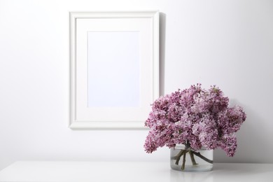 Photo of Beautiful lilac flowers in glass vase on table and picture frame indoors