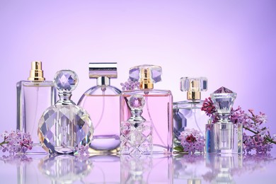 Photo of Bottles of perfume and lilac flowers on mirror surface against violet gradient background